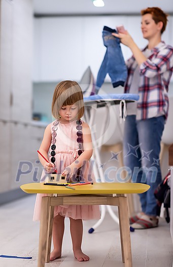 happy family spending time together at home cute little daughter in a pink dress playing and painting the jewelry box while young redhead mother ironing clothes behind her happy family spending time together at home cute little daughter in a pink dress playing and painting the jewelry box while young redhead mother ironing clothes behind her