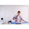 beautiful young red haired housewife in good mood ironing clothes on ironing board in front of a white wall at home