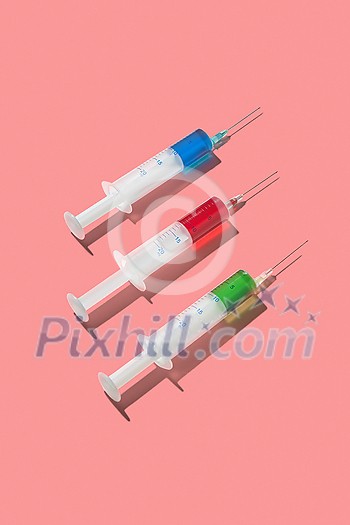 Three sterile disposable syringes 20 ml with red, green and blue serum or drugs for an intravenous injection and vaccinaction on a living coral background with hard shadow, copy space. Three sterile disposable syringes 20 ml with red, green and blue serum or drugs for an intravenous injection and vaccinaction on a living coral background with hard shadow, copy space.