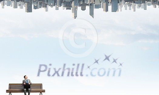 Young businessman sitting on bench with briefcase in hands and city reflecting in sky Young businessman sitting on bench with briefcase in hands and city reflecting in sky