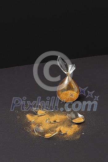 Brocken old-fashioned aged hourglass with bits of glass and golden sand on a duotone dark background with soft shadows, copy space. Brocken old-fashioned aged hourglass with bits of glass and golden sand on a duotone dark background with soft shadows, copy space.