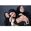 group of young beautiful muslim women in fashionable dress with hijab using mobile phone while taking selfie picture in front of black chalkboard representing modern islam fashion technology and ramadan kareem concept