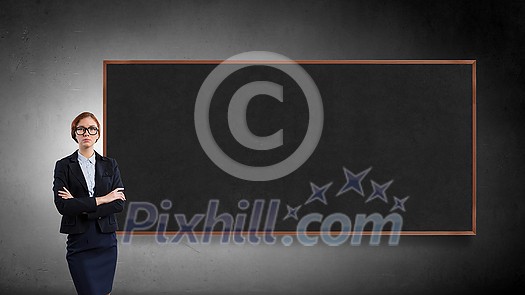 Young confident woman wearing glasses standing near blank blackboard Young confident woman wearing glasses standing near blank blackboard