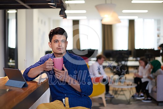 casual indian business man taking break from the work using tablet computer while drinking tea in relaxation area of modern open plan startup office casual indian business man taking break from the work using tablet computer while drinking tea in relaxation area of modern open plan startup office