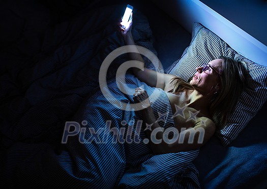 Pretty, middle-aged woman using her cell phone in bed at home before sleep Pretty, middle-aged woman using her cell phone in bed at home before sleep