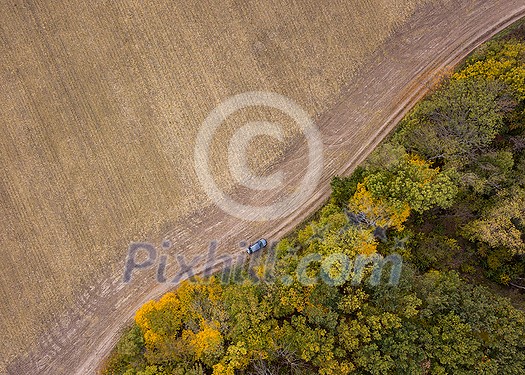 Top aerial panoramic view from a drone above dirt road across the agricultural field and moving car and forest area in an autumn season. Top aerial panoramic view from a drone above dirt road across the agricultural field and moving car and forest area in an autumn season.