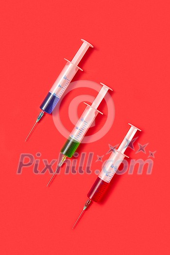Medical set of sterile plastic disposable syrenges 20 ml with colorful vaccines or serum for an intravenous injection on a red background with soft shadow, copy space. Top view. Medical set of sterile plastic disposable syrenges 20 ml with colorful vaccines or serum for an intravenous injection on a red background with soft shadow, copy space. Top view.