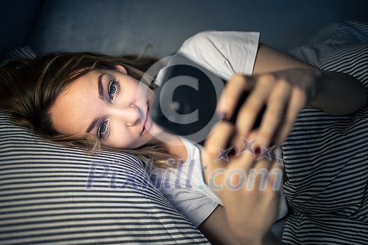 Young woman in bed holding a phone, tired and exhausted, blue light straining her eyes, messing up her circadian rhytm Young woman in bed holding a phone, tired and exhausted, blue light straining her eyes, messing up her circadian rhytm