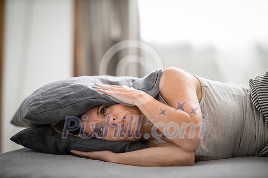 Sleepless lady covering ears with pillow. Noisy neighbors, tinnitus, insomnia or stress concept. Tired woman can't sleep. Awake in bed after coming home from her shift, work.