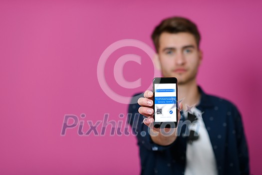covid passport, a young man shows his code and covid passport on his cell phone while posing in front of a pink background covid passport, a young man shows his code and covid passport on his cell phone while posing in front of a pink background