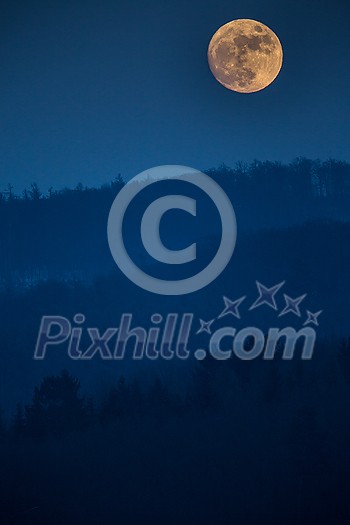 Atmospheric moon landscape of twilight in mountains. Dark lilac sky and big moon with cloud above silhouettes of mountain forests at dusk Atmospheric moon landscape of twilight in mountains. Dark lilac sky and big moon with cloud above silhouettes of mountain forests at dusk