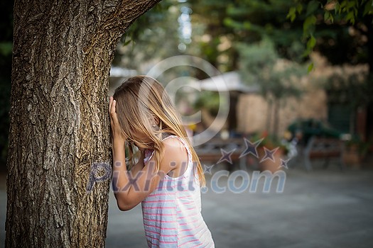 Little girl playing hide and seek with friends, counting by a tree, waiting for them to hide Little girl playing hide and seek with friends, counting by a tree, waiting for them to hide