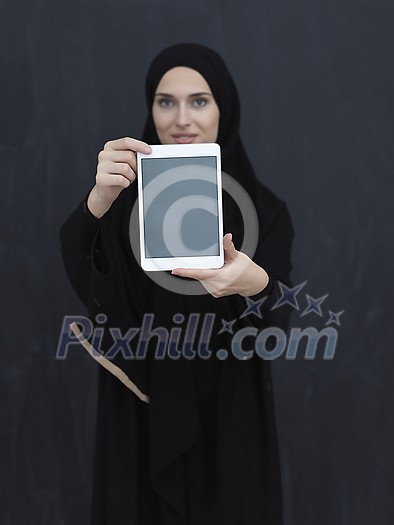 Young Arab businesswoman in traditional clothes or abaya and glasses showing tablet computer display in front of black chalkboard representing modern islam fashion and technology Young Arab businesswoman in traditional clothes or abaya and glasses showing tablet computer display in front of black chalkboard representing modern islam fashion and technology