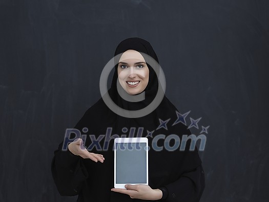 Young Arab businesswoman in traditional clothes or abaya and glasses showing tablet computer display in front of black chalkboard representing modern islam fashion and technology Young Arab businesswoman in traditional clothes or abaya and glasses showing tablet computer display in front of black chalkboard representing modern islam fashion and technology