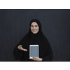 Young Arab businesswoman in traditional clothes or abaya and glasses showing tablet computer display in front of black chalkboard representing modern islam fashion and technology