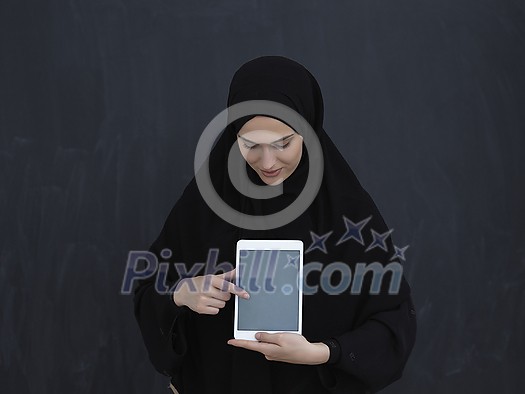 Young Arab businesswoman in traditional clothes or abaya and glasses showing tablet computer display in front of black chalkboard representing modern islam fashion and technology Young Arab businesswoman in traditional clothes or abaya and glasses showing tablet computer display in front of black chalkboard representing modern islam fashion and technology