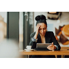 Young Latin woman with sad face sitting in the coffee shop at wooden table, drinking coffee and using a laptop.