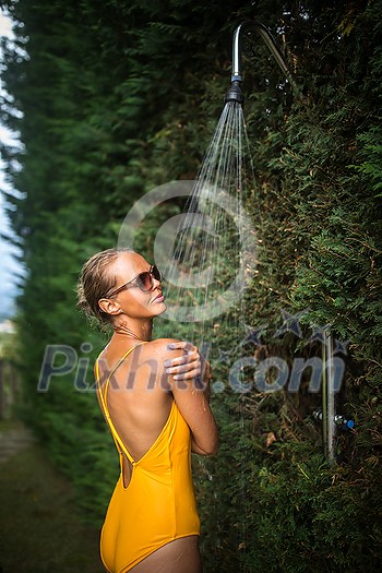 Beautiful young woman taking a shower outdoors. Attractive young woman in yellow one-piece enjoys shower on hot sunny day after a swim. Heat and refreshment concept. Beautiful young woman taking a shower outdoors. Attractive young woman in yellow one-piece enjoys shower on hot sunny day after a swim. Heat and refreshment concept.