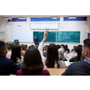 Rear view of female student sitting in the class and raising hand up to ask question during lecture. High school student raises hand and asks lecturer a question.