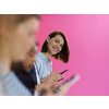 a group of diverse teenagers use mobile devices while posing for a studio photo in front of a pink background
