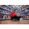 a young student with glasses sits in the library, reads a book and makes notes with a latpop