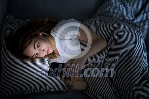 Young woman in bed holding a phone, tired and exhausted, blue light straining her eyes, messing up her circadian rhytm Young woman in bed holding a phone, tired and exhausted, blue light straining her eyes, messing up her circadian rhytm