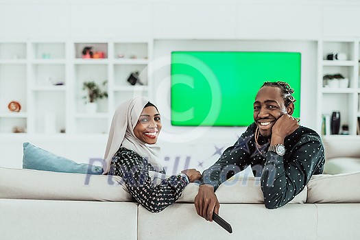 African Couple Sitting On Sofa Watching TV Together Chroma Green Screen Woman Wearing Islamic Hijab Clothes. High quality photo African Couple Sitting On Sofa Watching TV Together Chroma Green Screen Woman Wearing Islamic Hijab Clothes. High quality photo