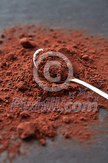 High angle of shiny spoon on heap of dry brown powder of Theobroma cocoa beans on gray surface High angle of shiny spoon on heap of dry brown powder of Theobroma cocoa beans on gray surface