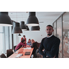 Portrait of happy Afro-American millennial male business owner in modern office. Businessman wearing glasses, smiling and looking at camera. Busy diverse team working in the background. Leadership concept. Head shot. High quality photo