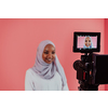 Videographer in digital studio recording video on professional camera by shooting female Muslim woman wearing hijab scarf plastic pink background. High-quality photo