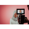 Videographer in digital studio recording video on professional camera by shooting female Muslim woman wearing hijab scarf plastic pink background. High-quality photo
