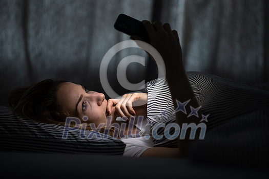 Young woman in bed holding a phone, tired and exhausted, blue light straining her eyes, messing up her circadian rhytm