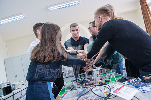 Group of young students in electronics classroom celebrating successfully finished project with holding their hands together, education and technology concept Group of young students in electronics classroom celebrating successfully finished project with holding their hands together, education and technology concept