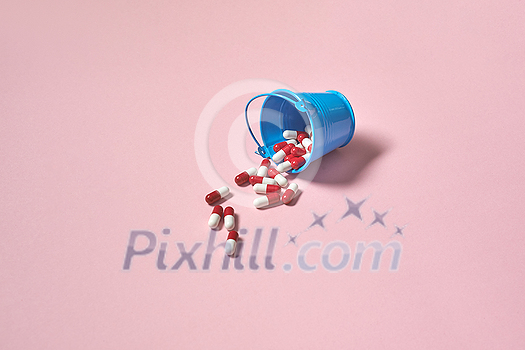 Pharmaceutical capsules falling out of bucket over pink background. Flat lay with copy space. Health care and medication-assisted treatment. Pharmaceutical capsules falling out of bucket over pink background. Flat lay with copy space. Health care and medication-assisted treatment.