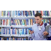 a young student with glasses stands in the school library with a smartphone, headphones and a laptop in his hand