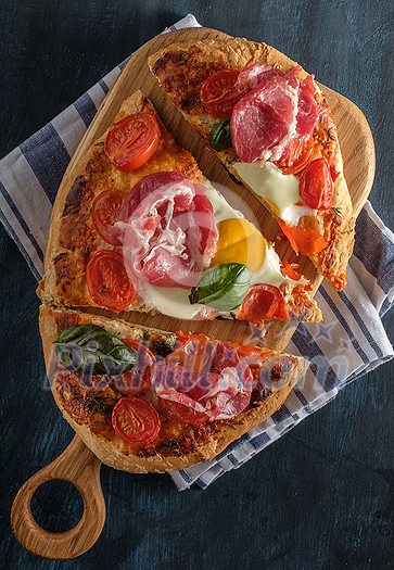 Sliced Homemade tasty pizza with bacon, egg, cheese, tomatoes and basil on dark background. Top view. Sliced Homemade tasty pizza with bacon, egg, cheese, tomatoes and basil on dark background. Top view.