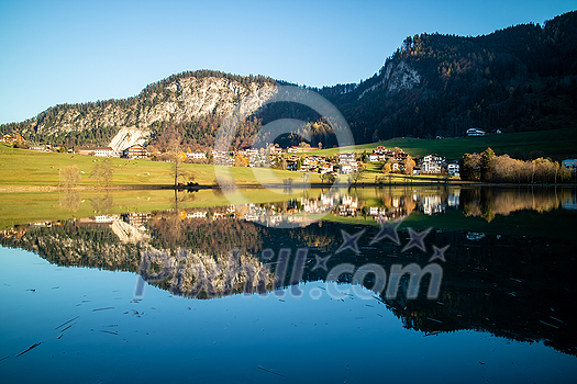 Amazing tranquil landscape with country area, houses and hills near the lake and reflection in clean blue water on a background of clear blue sky, Austria. Amazing tranquil landscape with country area, houses and hills near the lake and reflection in clean blue water on a background of clear blue sky, Austria.