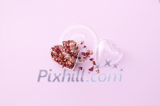 Dried red rose petals in open transparent heart-shaped pendant on toned background with space for text. Creative Valentine's day card design. Dried red rose petals in open transparent heart-shaped pendant on toned background with space for text. Creative Valentine's day card design.