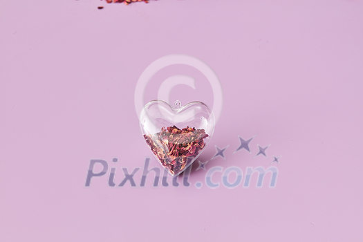 Red dried aroma rose petals inside small transparent heart-shaped pendant on toned background, close up with copy space. Creative wedding invitation card design. Red dried aroma rose petals inside small transparent heart-shaped pendant on toned background, close up with copy space. Creative wedding invitation card design.