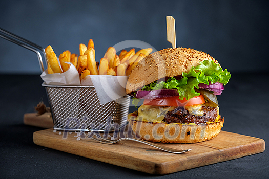 Tasty grilled beef burger with lettuce, cheese and onion served on cutting board with french fries on a black wooden table Tasty grilled beef burger with lettuce, cheese and onion served on cutting board with french fries on a black wooden table